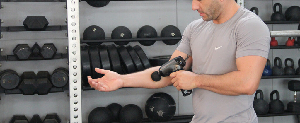Man using a Bob and Brad massage gun on his forearm in a gym