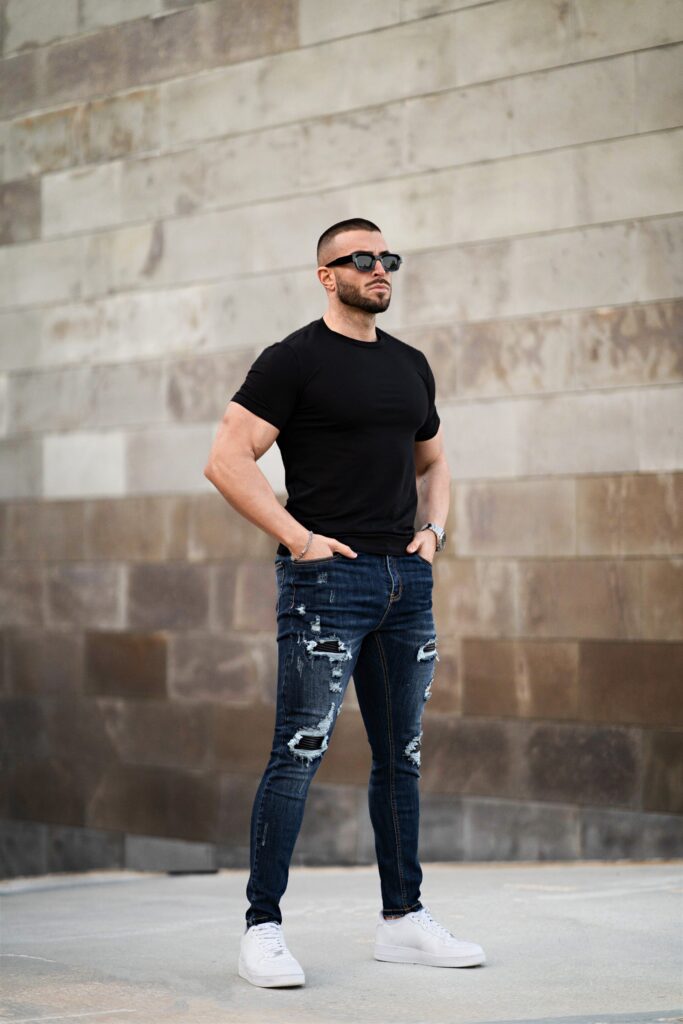 Three-quarter view of dark blue ripped skinny jeans with black patch details, worn with a black tee and white sneakers