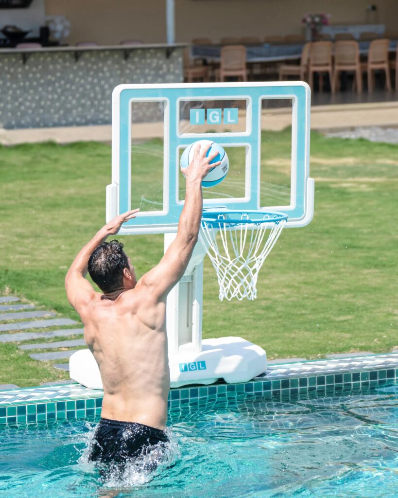 Swimmer reaching up to a ball at an IGL DunkSplash poolside basketball hoop with a teal backboard