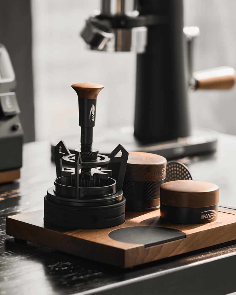 IKAPE espresso tools, including a tamper and distributors, on a walnut tray beside a grinder
