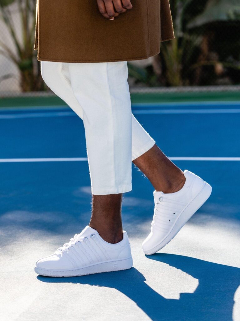Person in white trousers and white leather K-Swiss sneakers walking across a blue tennis court