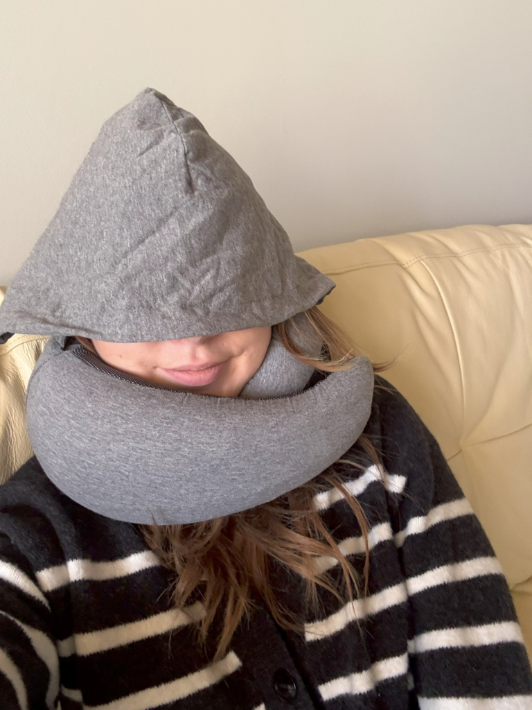 Person resting on a couch with the grey hood of a LoexAir Hooded Pro neck pillow pulled up over their eyes