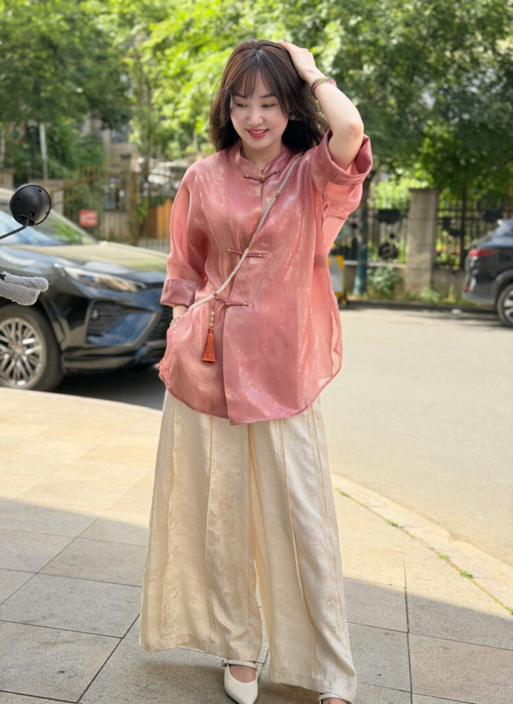 Model in the pink shimmer brocade blouse with frog-button closure, styled with wide cream trousers on a city street
