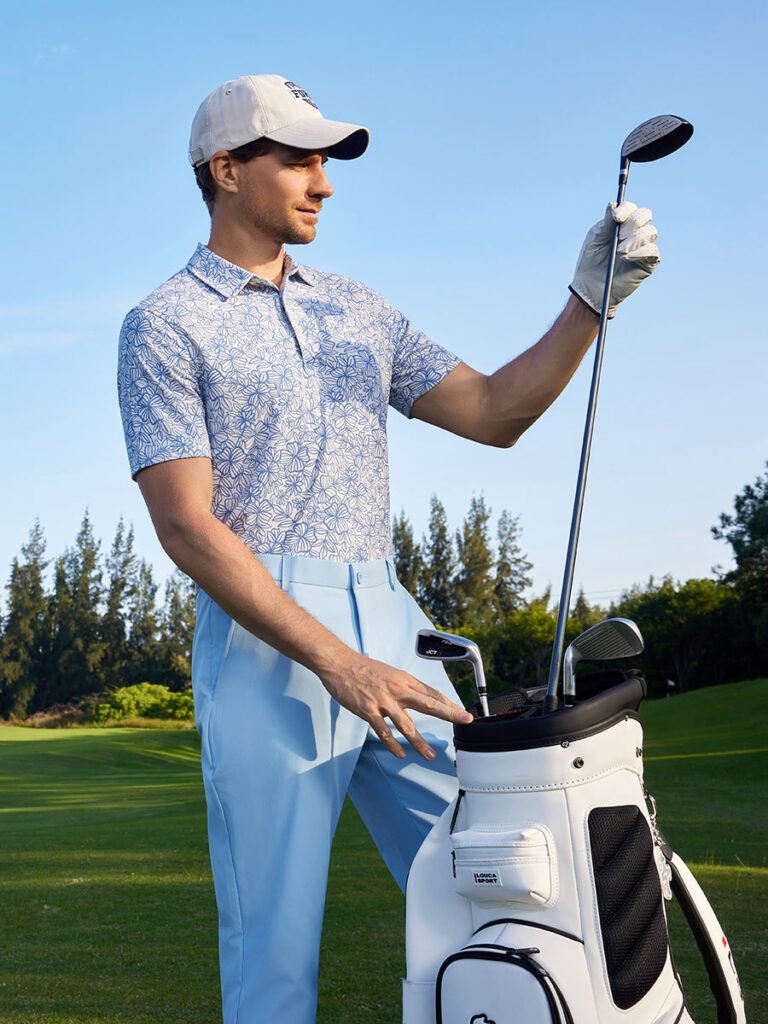 Man in a light blue floral-print golf polo and light blue pants reaching for a club in a white golf bag