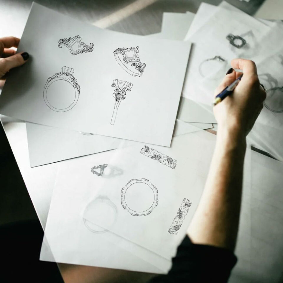 A designer's hands holding pencil sketches of leaf-and-vine ring designs over a desk of drawings