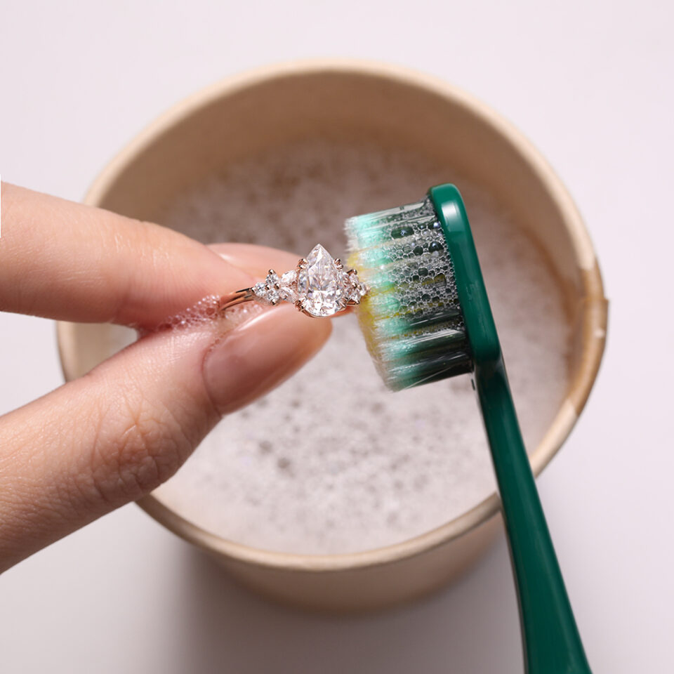 A pear-shaped moissanite ring being brushed with a soft toothbrush above a bowl of soapy water