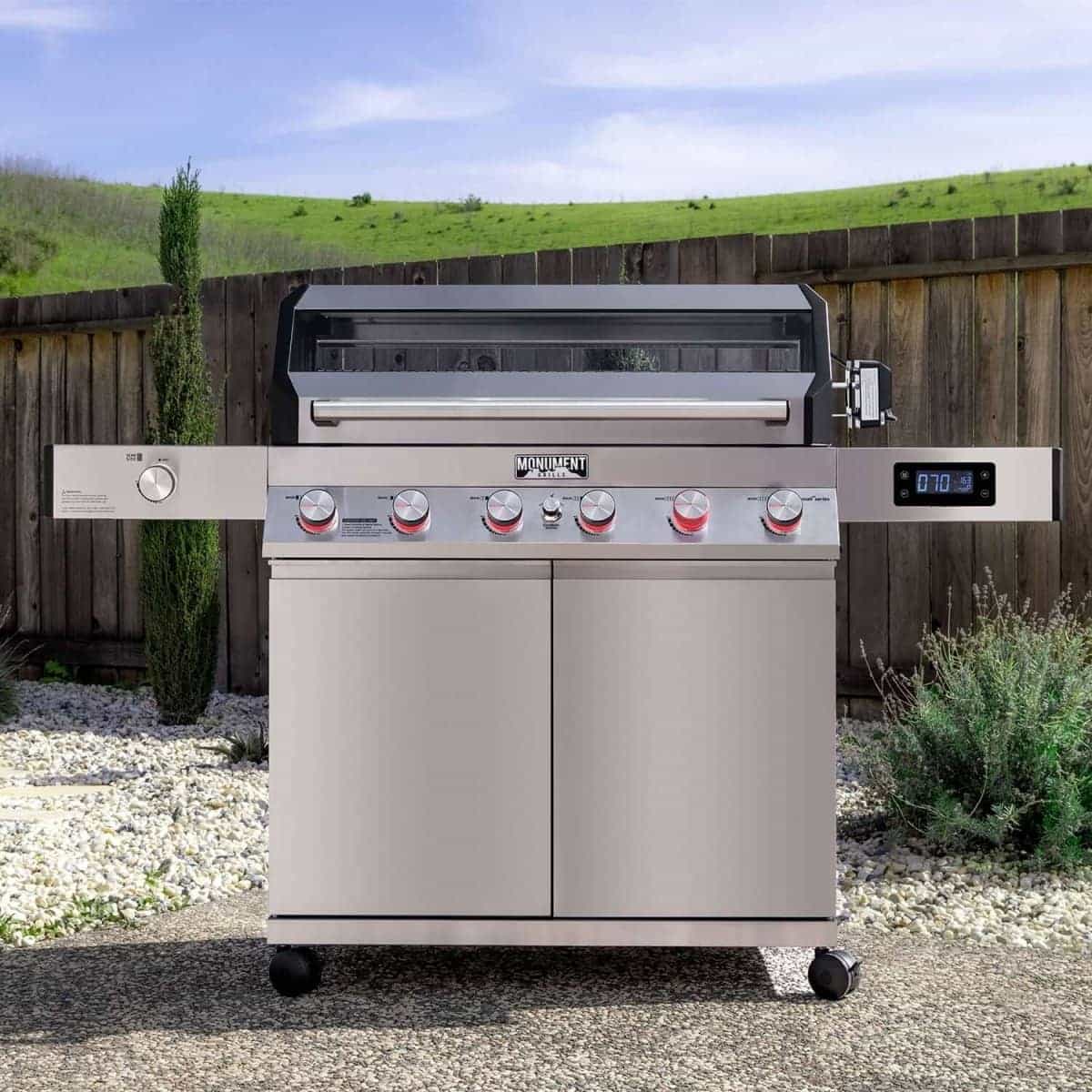 Stainless Monument Denali 605Pro gas grill with red-lit knobs in front of a wooden fence