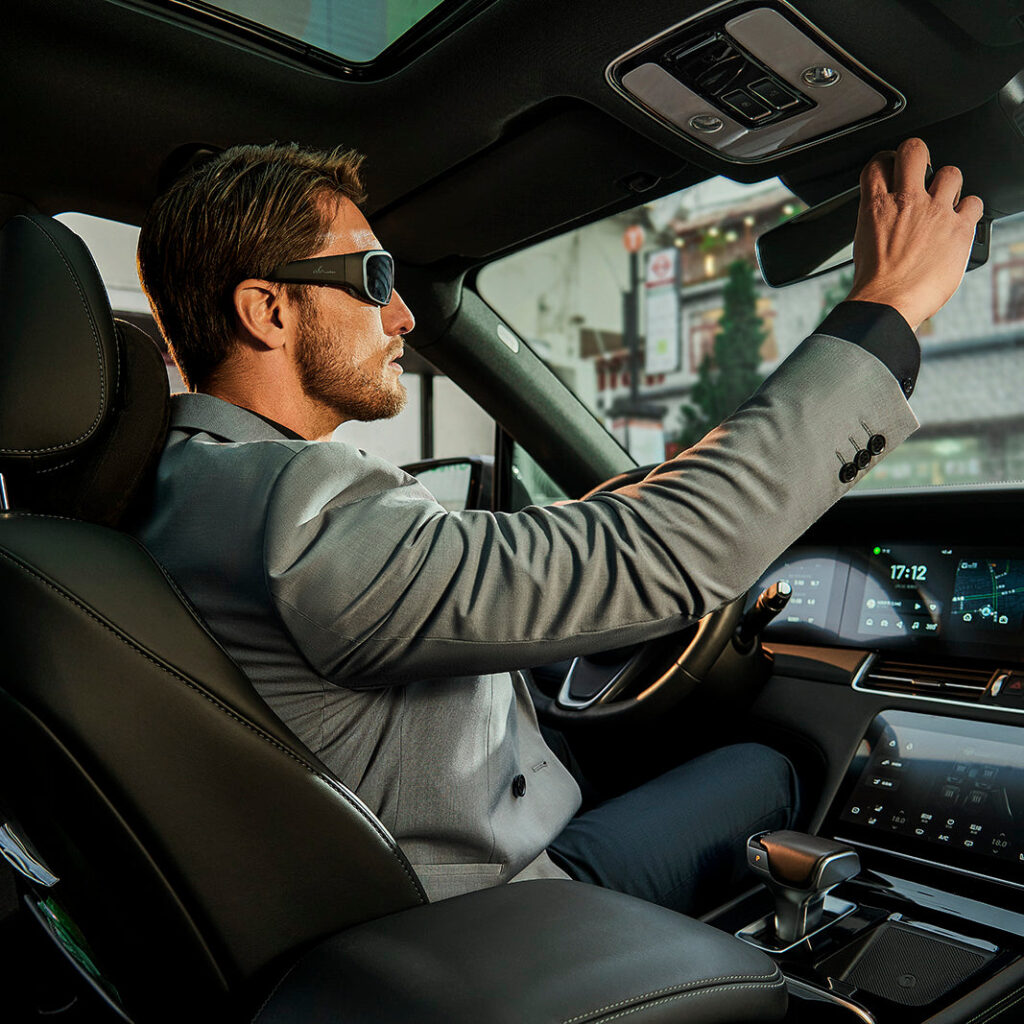 Man in a gray blazer wearing black wraparound Bluetooth sunglasses while adjusting the rearview mirror inside a car