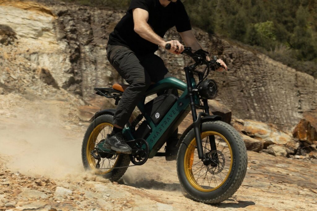Rider leaning a green Smartravel fat-tire e-bike through a dusty trail corner beside a rock wall