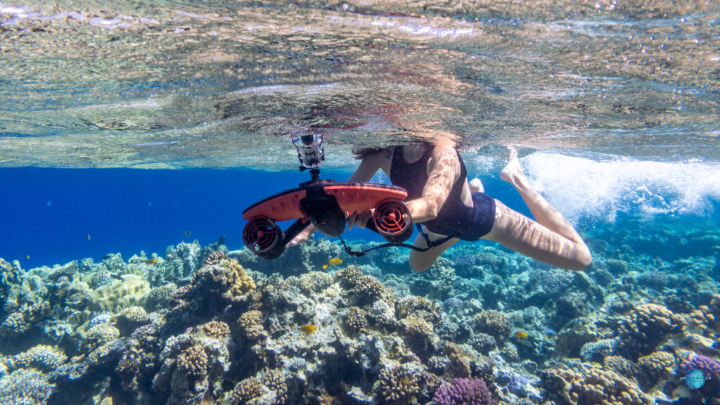 Swimmer holding a red SUBLUE underwater scooter with an action camera mounted on top, above a coral reef
