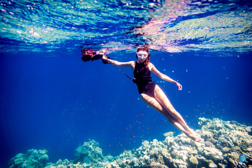 Woman in a swimsuit holding a SUBLUE underwater scooter in clear blue water above a coral reef