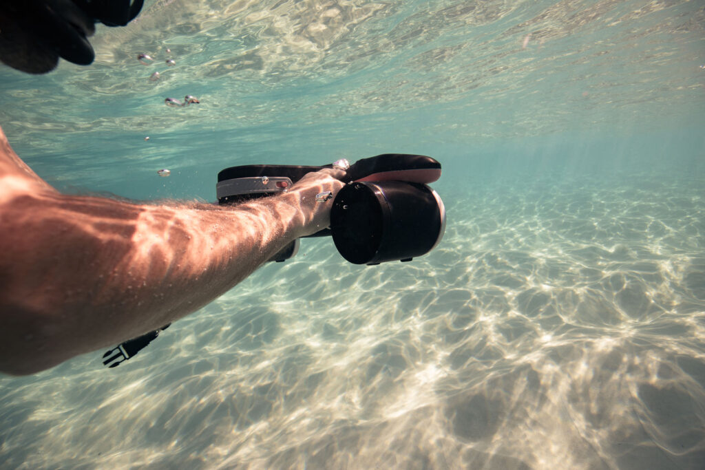 Swimmer's arm holding a SUBLUE underwater scooter in shallow, sunlit water over a sandy seabed