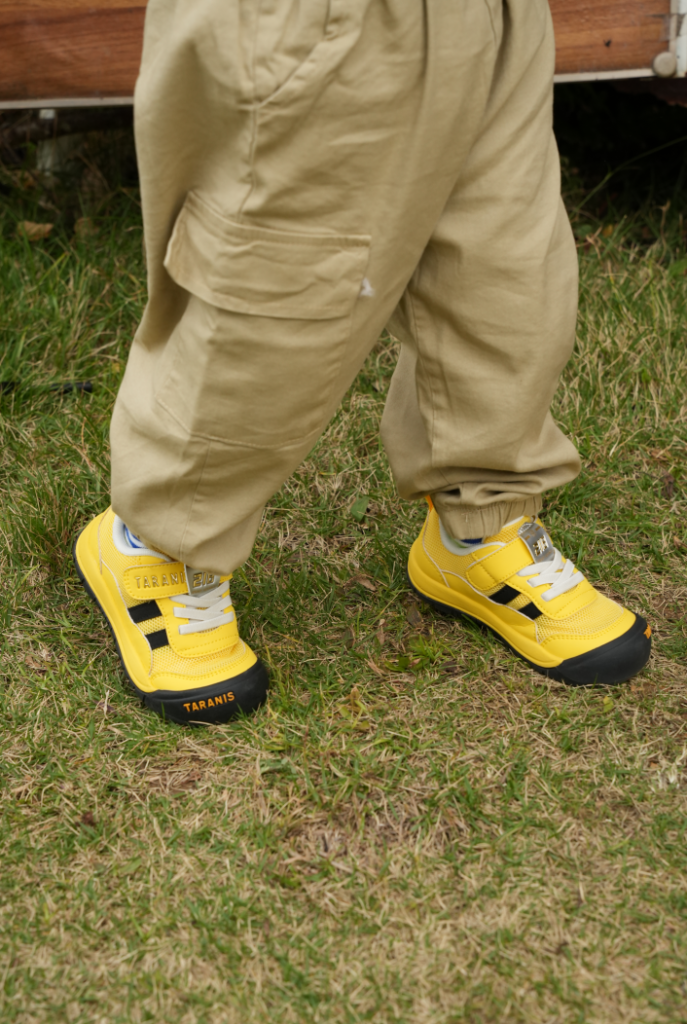 Toddler standing on grass in yellow Taranis shoes with black toe caps, wearing khaki cargo trousers