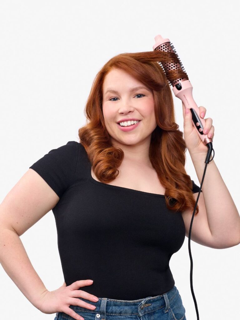 A smiling woman with wavy red hair styling a section with a pink Wavytalk thermal brush