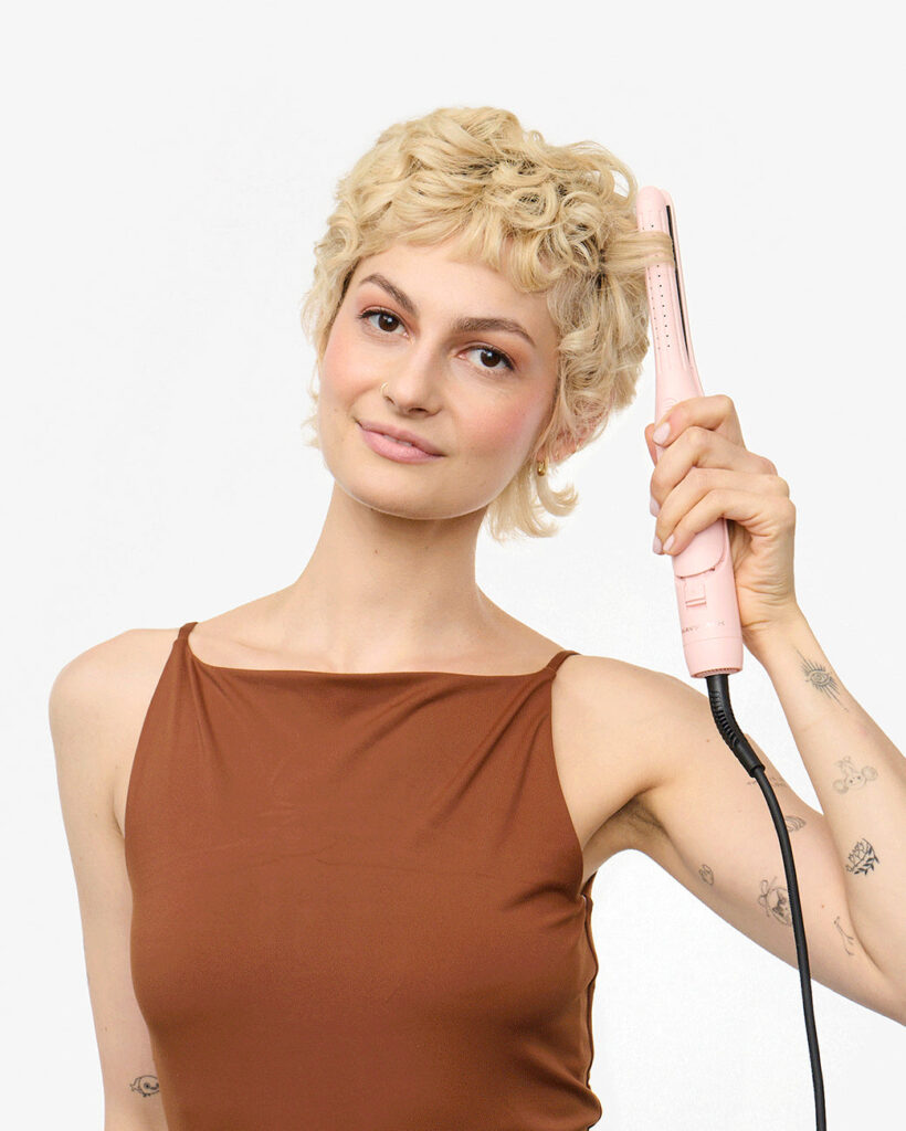 A woman with short blonde curls wrapping a section of hair around a slim pink Wavytalk curling iron