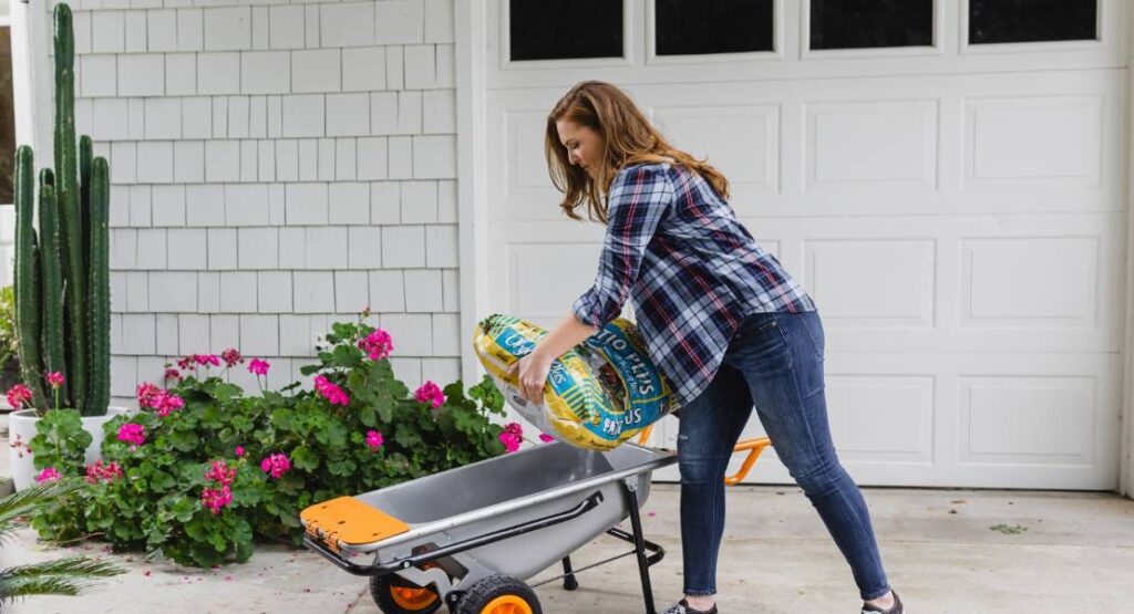 Woman tipping a bag of potting soil into a WORX Aerocart wheelbarrow beside a flower bed