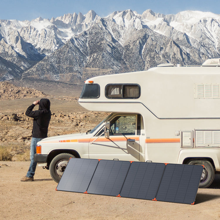 Large foldable solar panel with orange corners set up on the ground beside a camper, with snowy mountains behind